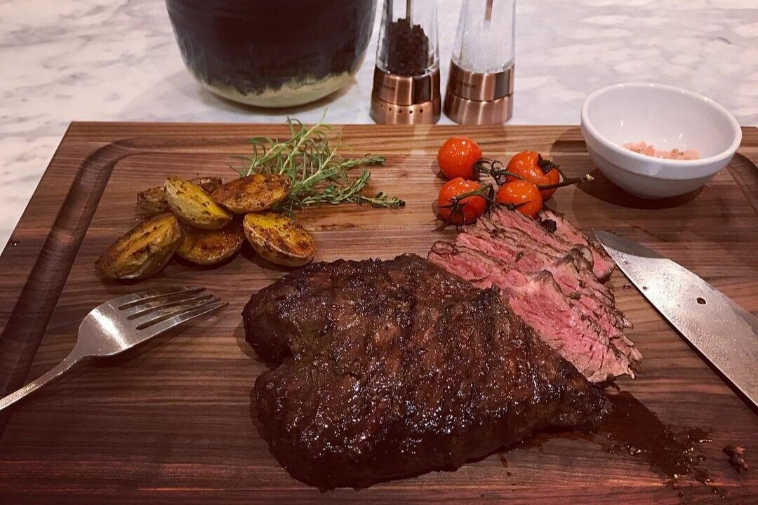 Grilled steak served on a wooden cutting board, accompanied by roasted potatoes, cherry tomatoes, fresh thyme, and a bowl of pink salt, with a fork and knife nearby for serving.