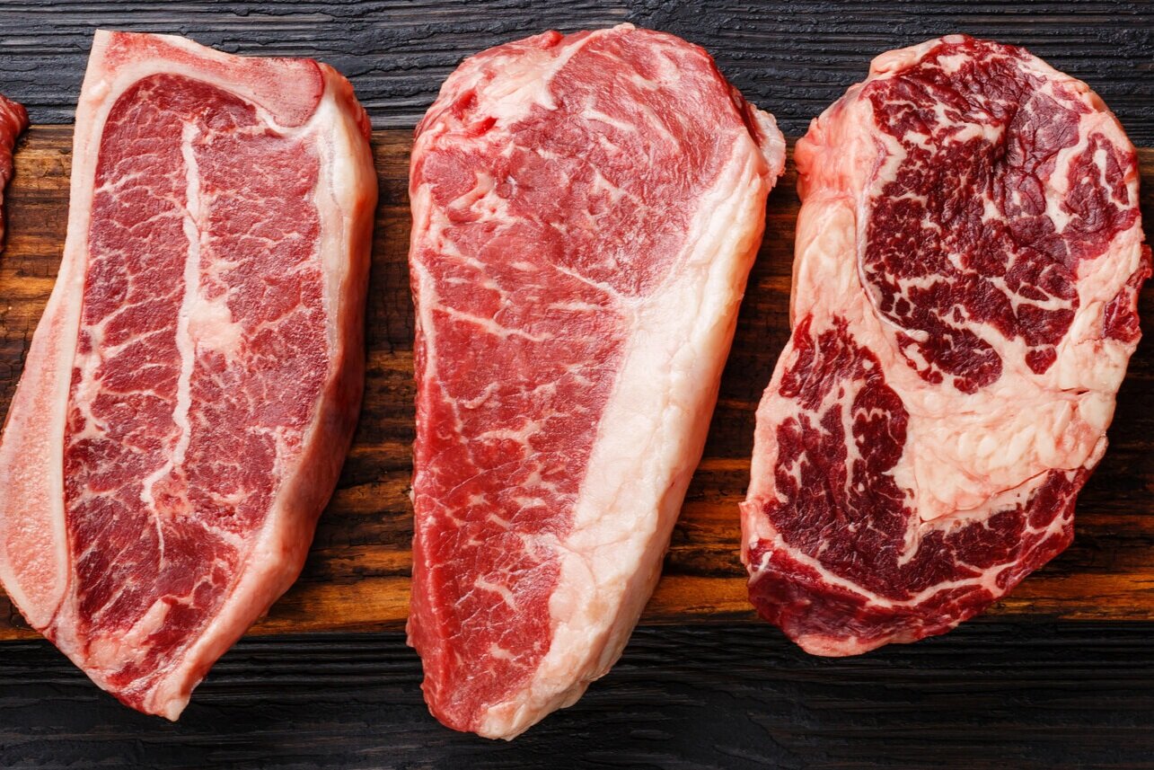 Assorted cuts of raw steak displayed on a wooden cutting board, showcasing marbling and texture differences, ideal for grilling or culinary preparation.