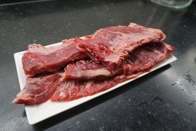Raw beef flank steak displayed on a white platter, showcasing its rich red color and marbling, set against a dark countertop background. Ideal for grilling or slow cooking recipes.